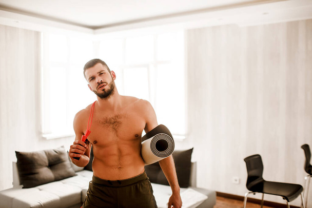 Jeune homme faisant de l'exercice à la maison sur le canapé. Il tient un tapis de yoga et une corde à sauter et regarde la caméra.
 - Photo, image
