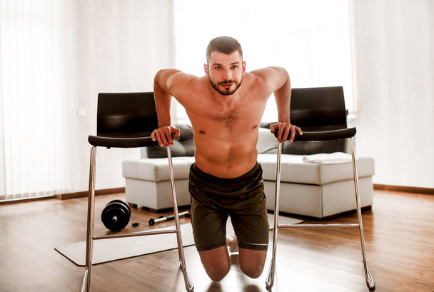 Young man doing sport workout in room during quarantine. T-shirtless guy sportsman doing workout at home He is trains triceps with chairs - Foto, afbeelding