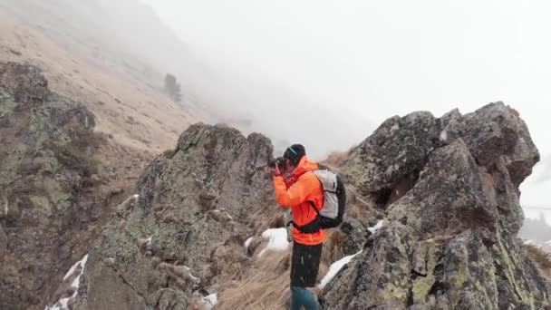 Aerial view of a young male photographer with a camera in his hands stands on a high rock in a gorge and photographs an alpine waterfall during a snowstorm and snowfall in spring. - Footage, Video
