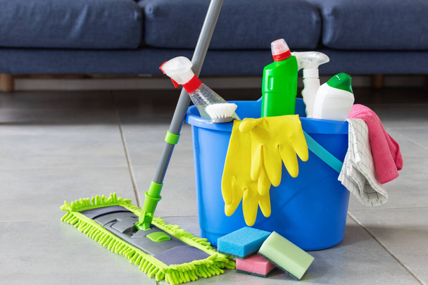 Bucket with sponges, bottles of detergent and mopping stick in front of sofa in living room. Housework cleaning equipment. Cleaning service idea. - Zdjęcie, obraz