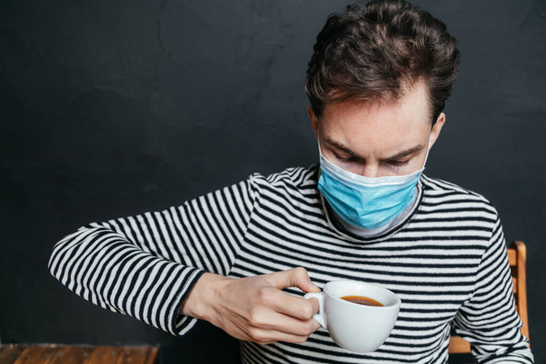 Portrait of an attractive caucasian man with surgical medical face mask who is concerned about preventing pandemic coronavirus on a black background.  Coronavirus Covid-19 concept. Protect your health. Drinking coffee - Zdjęcie, obraz