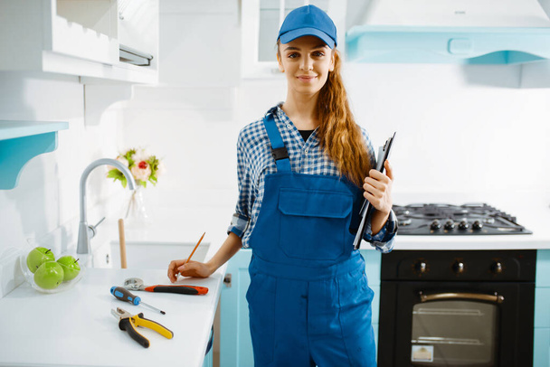 Cute female furniture maker in uniform holds notebook in the kitchen. Handywoman fixing problem with garniture cabinet, repairing service at home - Foto, Imagen