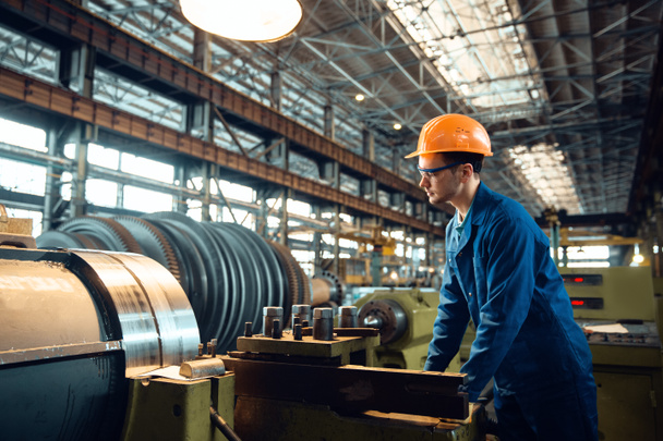 Male worker in uniform and helmet checks turning lathe on plant. Industrial production, metalwork engineering, power machines manufacturing - Photo, Image