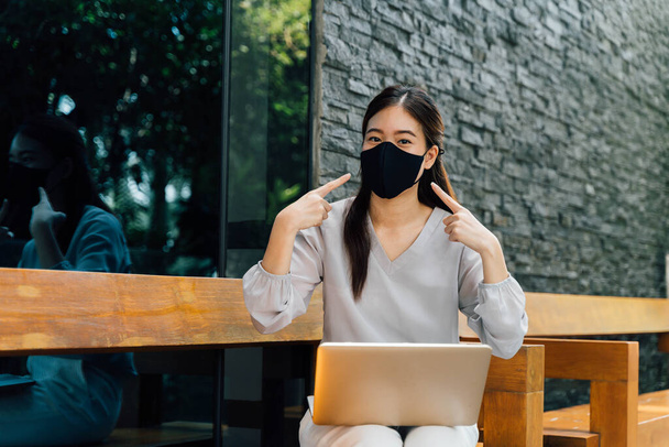 Asian woman wearing a face mask outside while sitting on laptop and pointin at her mask. Dont forget to wear a mask and face mask requirement to go outside concept - Foto, afbeelding