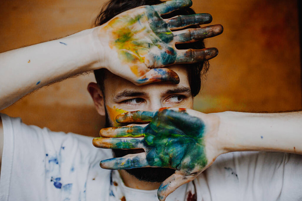 Portrait of a young creative artist, man wearing a white T-shirt with colored spots from paint posing for a photo in the process of creating a picture - Fotó, kép