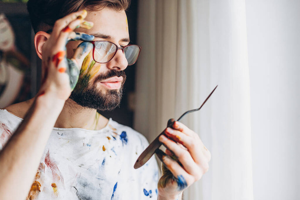 Portrait of a young creative artist, man wearing a white T-shirt with colored spots from paint posing for a photo in the process of creating a picture - Foto, Imagem