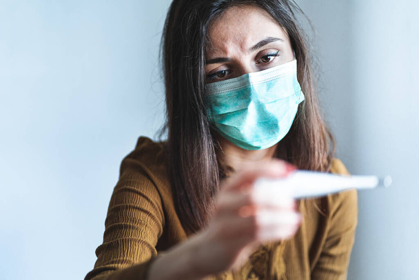 Sick young woman wearing a surgical mask and holding a digital thermometer that indicates she has over 38 degrees fever. Sick and worried female with fever and illness during pandemic. Quarantine, self isolation and coronavirus concept. Headache. Flu - Foto, Imagem