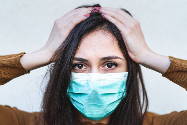 Close up portrait of disturbed annoyed young woman holding hands above head wearing a surgical mask feeling sick and worried about epidemic outbreak. Anxious lady touching head with stress migraine feeling depressed in quarantine. - Photo, image