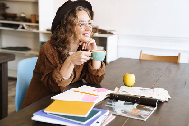 Image of joyful charming woman in eyeglasses drinking coffee while studying with exercise books at home - 写真・画像