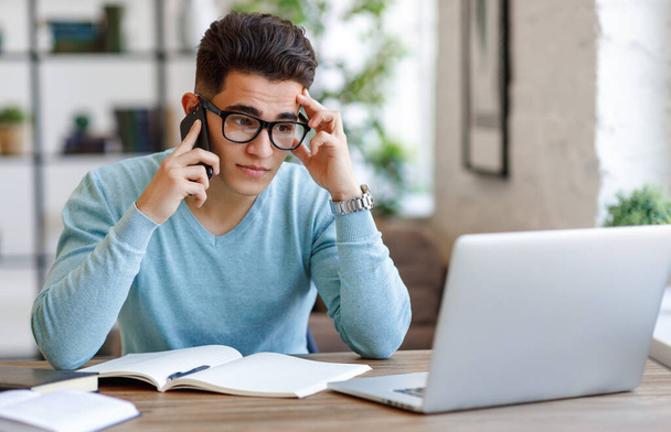 Κουρασμένος νεαρός άνδρας με γυαλιά που αγγίζει το μέτωπο και διαβάζει δεδομένα από το laptop ενώ μιλάει σε smartphone κατά τη διάρκεια εργασιών σε απομακρυσμένο έργο στο hom - Φωτογραφία, εικόνα