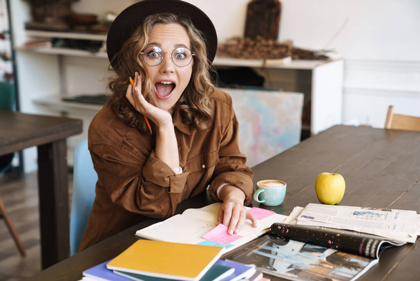 Image of attractive woman in eyeglasses and hat expressing surprise while studying with exercise books at home - Zdjęcie, obraz