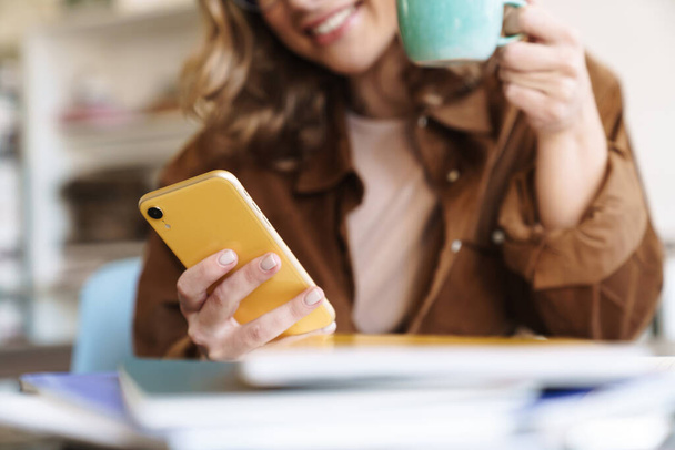 Image of smiling nice woman in eyeglasses and hat using cellphone while drinking coffee at home - Photo, Image