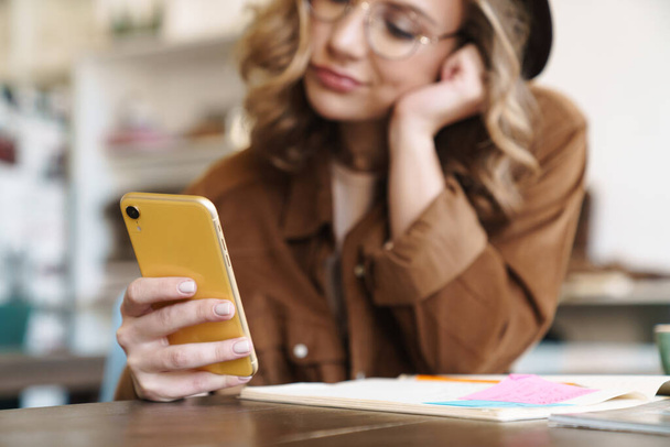 Image of displeased nice woman in eyeglasses and hat using cellphone while studying at home - Foto, Imagem