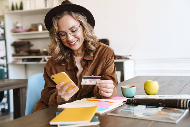 Image of smiling nice woman in hat using cellphone and holding credit card while studying at home - Foto, Imagem