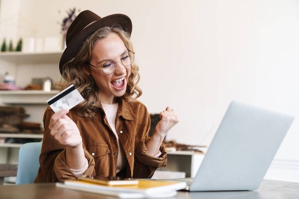 Image of delighted young woman in hat making winner gesture and holding credit card while working with laptop - Foto, Bild