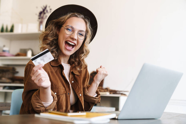 Image of delighted young woman in hat making winner gesture and holding credit card while working with laptop - Foto, Imagem