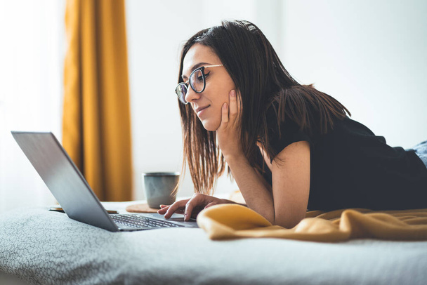 Улыбающаяся привлекательная женщина лежит на кровати с помощью ноутбука, общаясь онлайн дома, счастливая девушка в очках и печатая на компьютере, наслаждаясь покупками в Интернете или чате в социальных сетях
 - Фото, изображение