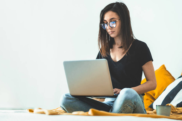 Atrakcyjna młoda kobieta w okularach, pracująca w domu. Kobieta przedsiębiorca siedzi w łóżku i pracuje na laptopie pijąc poranną kawę. Uczenie się do egzaminu. - Zdjęcie, obraz