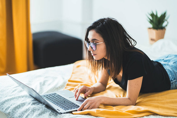 Pretty brunette woman wearing eyeglasses and working on laptop computer. Concentrated female lying in bed at home writing and using notebook keyboard touchpad. Freelance lifestyle and work from home concept - Photo, Image