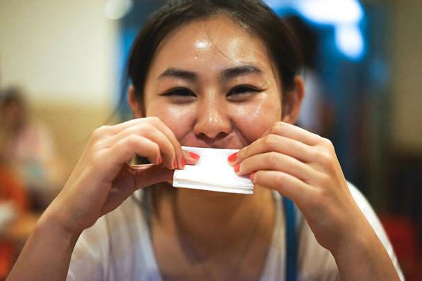 Nuori onnellinen hymyilevä aasialainen tyttö pyyhkii suun syötyään ruokaa ravintolassa, valkoista kudosta, hyvää tapaa pöydässä, onnea, valikoivaa keskittymistä, hämärtynyt bokeh, tumma kalvo melua viljaa - Valokuva, kuva