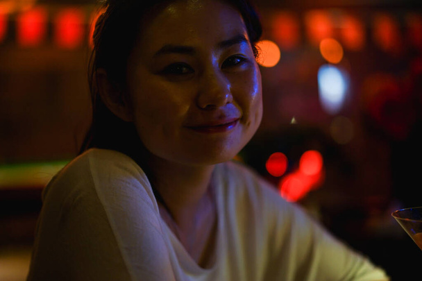 Genç ve mutlu Asyalı bir kadının portresi, karanlık bir gece barında, fotokopi makinesinde, randevuda olan bekar bir kız, bulanık arka plan, sahte ışıklar. - Fotoğraf, Görsel