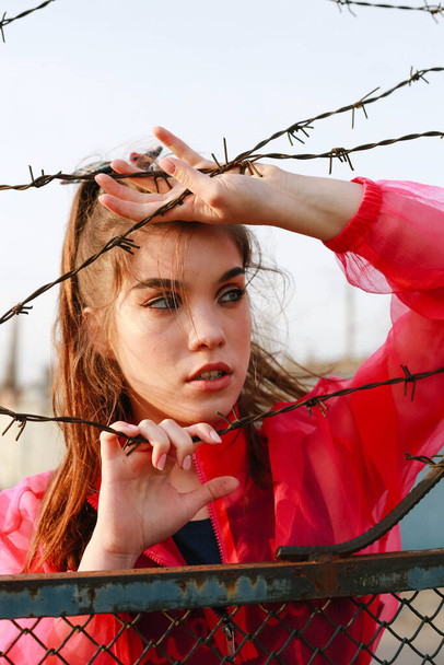 The girl behind the barbed wire holding a wire looks away on sun in better future - 写真・画像