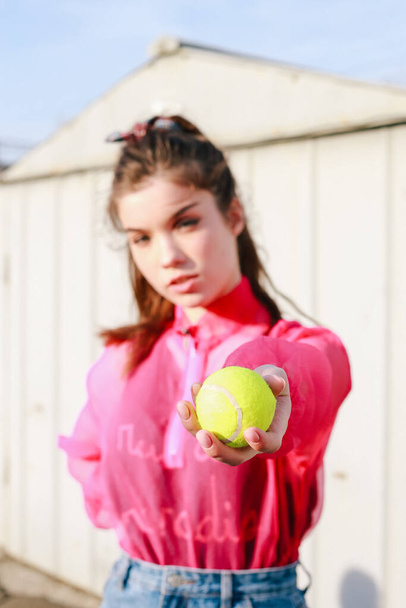 Blurred trendy girl in pink blouse give yellow tennis ball in front posing in garages. Street style portrait - Foto, Imagen