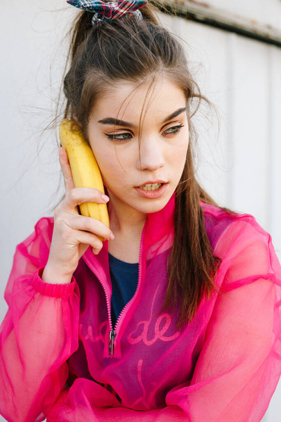 Model girl talking on banana in pink blouse posing in garages. Street style trendy portrait - Fotó, kép
