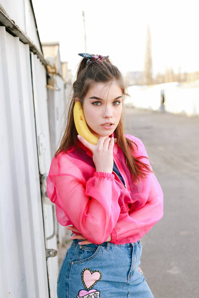 Model girl talking on banana in pink blouse posing in garages. Street style trendy portrait - Photo, Image
