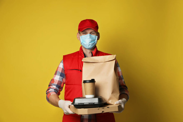 Courier in protective mask and gloves holding order on yellow background. Food delivery service during coronavirus quarantine - Fotó, kép