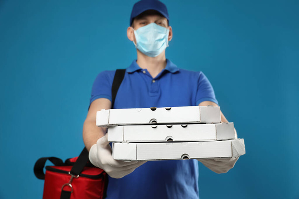 Courier in protective mask and gloves holding pizza boxes on blue background. Food delivery service during coronavirus quarantine - 写真・画像