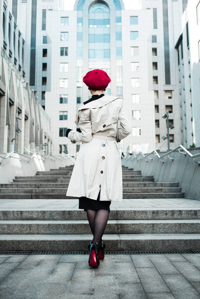 Un dorso di giovane eleganza ragazza con berretto rosso in mantello leggero e abito nero passeggiando sulle scale vicino al centro commerciale sulla città. Giornata di sole primaverile. Donna d'affari. Stile di vita
.  - Foto, immagini