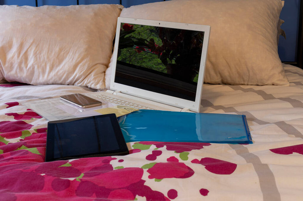 Вещи для работы из дома, лежащие на кровати. Компьютер, мобильный телефон, планшет и некоторые документы, готовящиеся к работе в постели
. - Фото, изображение
