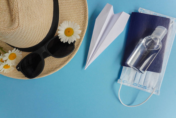Diseño de verano con sombrero de paja, margaritas y gafas de sol, avión-origami, desinfectante, pasaporte y máscara médica.Concepto de viaje y recreación. Vuelos al mar, descanso durante coronavirus. Pisos en azul
 - Foto, Imagen