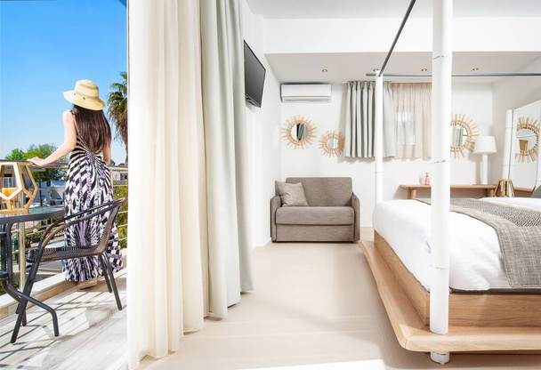 モダンなリゾートホテルのベッドルームのバルコニーに立つロマンチックなスタイルのドレスの若い女性。白、グレー、木製の家具と光のオープンスペースの部屋のインテリア - 写真・画像