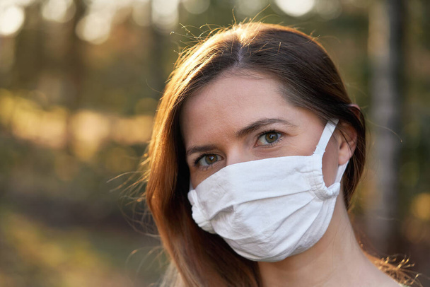 Young woman wearing white cotton virus mouth nose mask, blurred sunset lit trees in background, closeup face portrait - Фото, зображення
