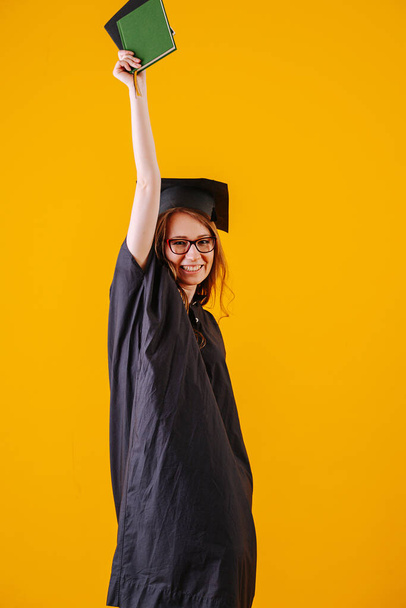 Woman celebrating her graduation, wearing gown and hat over orange background. Throwing hand with diploma in the air. - Φωτογραφία, εικόνα