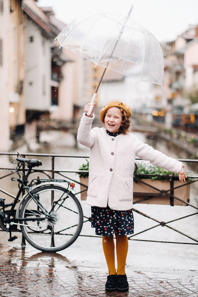 cheerful beautiful girl in a coat with a transparent umbrella in Annecy. France. The girl Cheerfully raises an umbrella in the rain - Photo, Image
