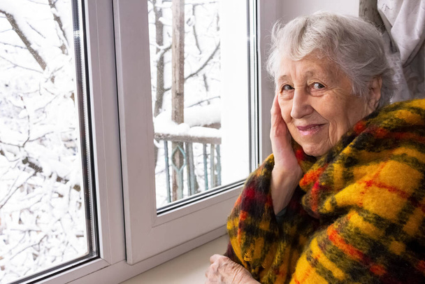 Vecchia donna sola seduta vicino alla finestra nella sua casa e guardando la fotocamera e sorridendo all'inverno - Foto, immagini