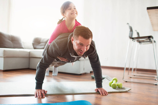 father and daughter are training at home. Workout in the apartment. Sports in home conditions. They make the doing plank. daughter climbed on dad. He is pushing up from the floor. - 写真・画像