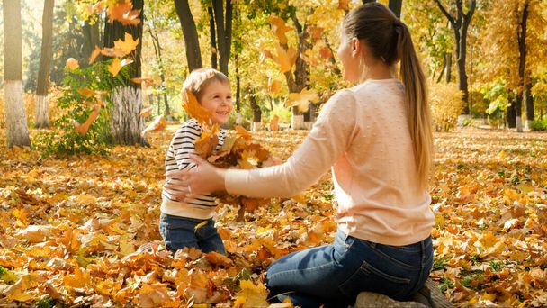 Happy laughing boy with young mother sitting on ground at park and throwing up autumn leaves - Foto, immagini