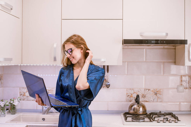 Smiling young woman holding blue laptop computer in hands and touching hair.Girl in blue bathrobe standing in modern light kitchen interior indoor.Shopping online,order food by internet,freelancing. - Photo, Image