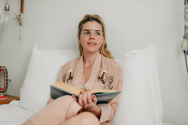 Ambiente domestico accogliente. Beatiful modello femminile indossa pigiami, si siede su lenzuola in camera da letto, libro, lavoro dal concetto di casa
 - Foto, immagini