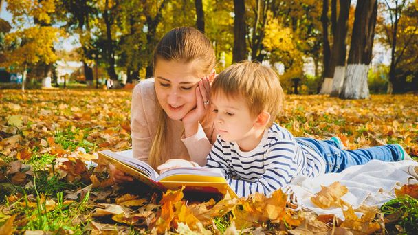 Genç annenin sonbahar yapraklarının üzerinde yatan oğluna kitap okurken çekilmiş tonlarca portresi. - Fotoğraf, Görsel