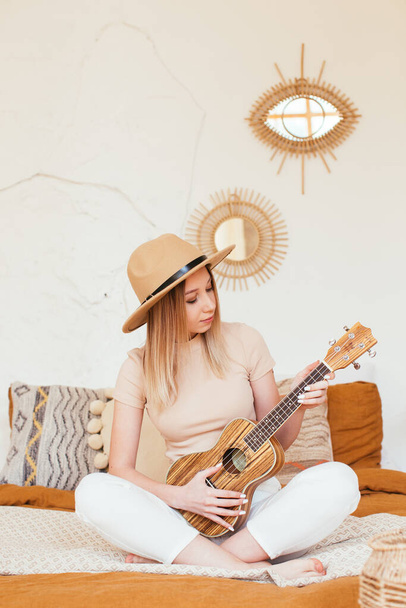 Young funny hipster girl having fun and playing on small ukulele guitar, singing and dancing. wearing a hat, joy, positive mood. instagram live stream concert concept - Foto, afbeelding