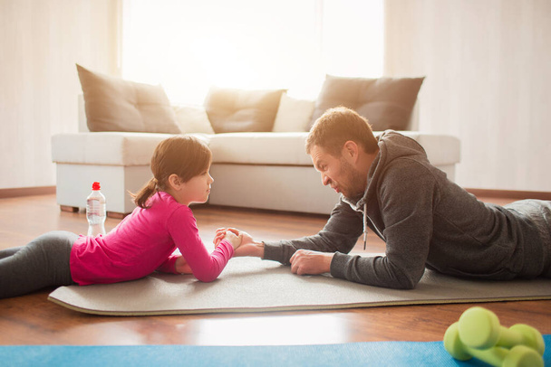 father and daughter are training at home. Workout in the apartment. Sports at home. Compete in arm wrestling and lie on a yoga mat - Zdjęcie, obraz