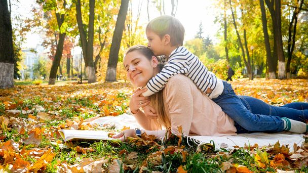 Kleines Kleinkind sitzt auf dem Rücken der Mutter und küsst sie im Herbstpark - Foto, Bild