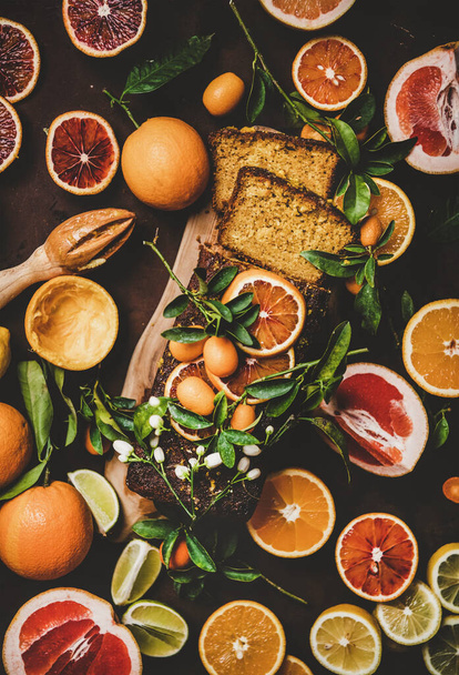 Citrus Mediterranean loaf cake. Flat-lay of almond pound cake with oranges, lemons, kumquats, grapefruits and blossom flowers over dark rusty background, top view. Flourless, vegan dessert concept - Foto, imagen