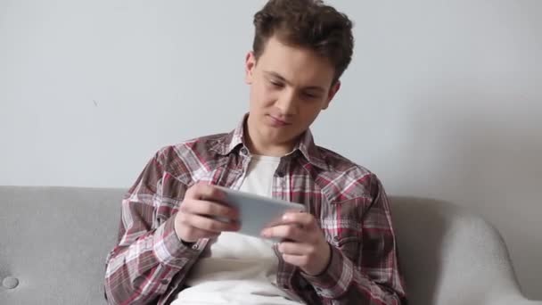 Student guy playing games on smartphone sitting in a living room on the couch. Young guy relaxing while gaming before or after studies. Prores 422 - Materiał filmowy, wideo