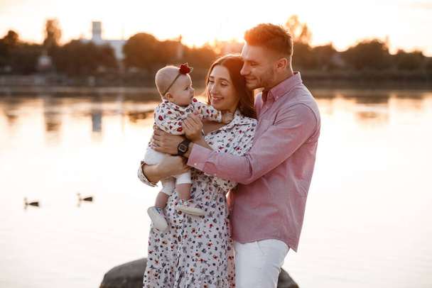 Happy young family near lake, pond. Family enjoying life together at sunset. People having fun in nature. Family look. Mother, father, child smiling while spending free time outdoors - Zdjęcie, obraz